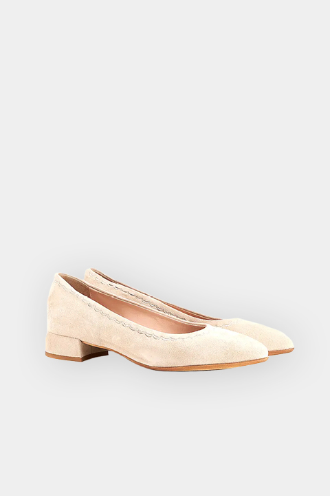 Un par de zapatos de salón de ante beige con punta afilada, bordes festoneados y suelas marrones, representados sobre un fondo gris claro.
