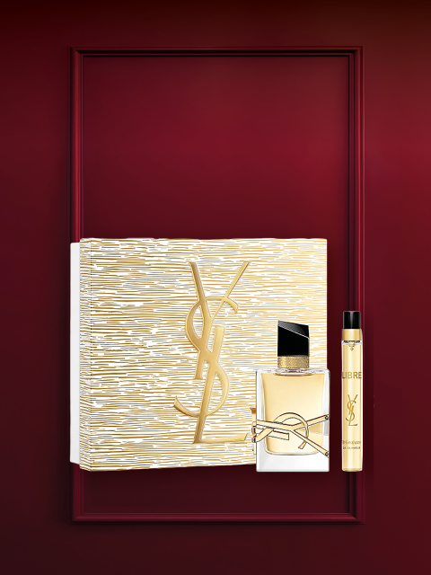 Set de regalo de fragancia en un marco rojo oscuro con una caja con diseño floral, un frasco pequeño de perfume y un tubo blanco con tapa dorada. En la parte superior, en letras blancas, se lee "Sets de fragancia".