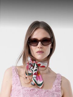 Mujer con un vestido rosa texturizado con motivos de rosetas, con gafas de sol oscuras y un pañuelo colorido atado al cuello, de pie frente a un fondo sencillo.