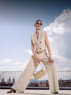 Una mujer con un traje pantalón beige sin mangas y gafas de sol posa al aire libre, con un bolso a juego en la mano, con un paisaje urbano y un cielo azul de fondo.