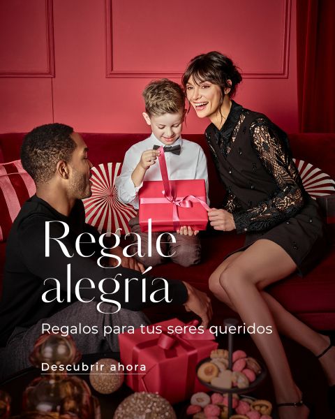 Una mujer, un hombre y un niño pequeño están sentados en un sofá rojo. El niño, sonriendo, está desenvolviendo un paquete de regalo rosa, mientras los adultos observan. La habitación está decorada festivamente en rojo. El texto en español dice: "Regala alegría. Regalos para tus seres queridos."