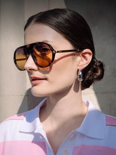 Una mujer con cabello castaño recogido en un moño bajo lleva unas grandes gafas de sol color ámbar, pendientes plateados y una camisa rosa y blanca con cuello, expuesta a la luz del sol.