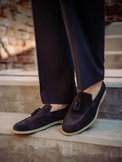 Persona con zapatos de gamuza oscura con borlas y pantalones oscuros, de pie en una escalera de concreto al aire libre con un fondo borroso de pared de ladrillo.