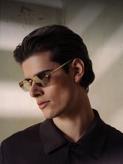 Un joven con cabello oscuro, gafas de sol marrones y camisa oscura con cuello mira hacia un lado con una luz suave y sombreada.
