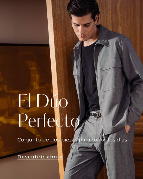 Un hombre con un elegante traje de dos piezas gris y una camisa negra está de pie en una casa junto a una pared de madera. Un texto en español promociona el atuendo.