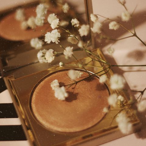 Un iluminador dorado y compacto con pequeñas flores blancas en la punta, que se reflejan en un espejo, creando una estética suave y elegante.