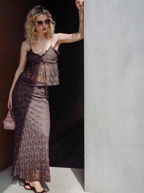 Una mujer con cabello rubio rizado, que lleva gafas de sol y un vestido de encaje marrón, se apoya contra una pared bajo la luz del sol brillante, con un bolso pequeño en la mano.