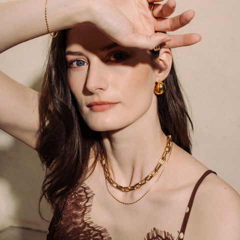 Mujer de piel clara que lleva joyas de oro y un top de encaje marrón y posa con un brazo levantado para proteger parcialmente su rostro de la luz.
