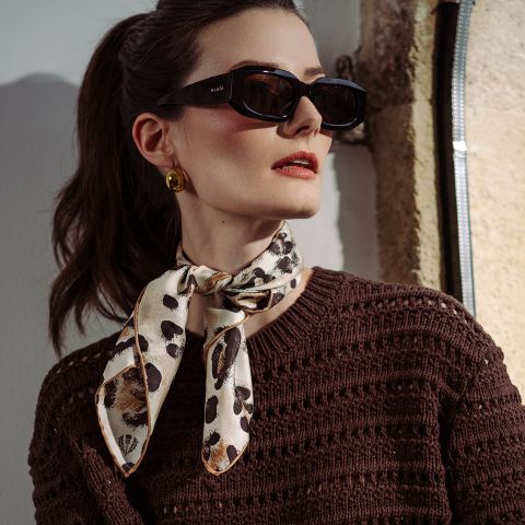 Mujer con gafas de sol negras, pendientes dorados, un top de punto marrón y una bufanda estampada atada al cuello, de pie bajo la luz del sol.