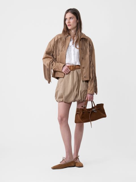 Mujer con chaqueta de flecos marrón claro, blusa blanca, pantalones cortos marrón claro con cinturón, zapatos planos marrones y un bolso marrón frente a un fondo sencillo.