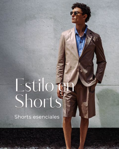 Hombre con chaqueta de traje color topo y pantalones cortos a juego, con camisa de rayas azules, gafas de sol y una mano en el bolsillo. El texto dice "Estilo en Shorts.".