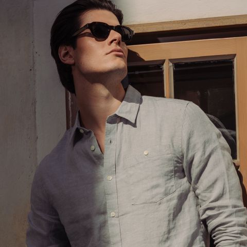 Un hombre con gafas de sol y una camisa abotonada gris claro está de pie junto a la ventana y mira al sol.
