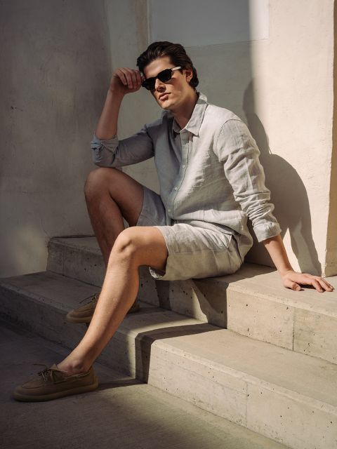 Un hombre con gafas de sol, camisa de lino clara y pantalones cortos está sentado en una escalera de hormigón a la luz del sol y apoya un brazo en su rodilla.