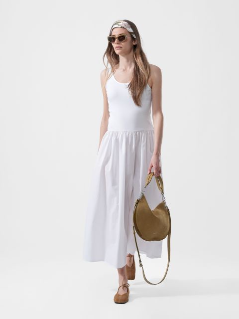 Mujer con un vestido maxi blanco sin mangas, gafas de sol, pañuelo en la cabeza, bailarinas marrones claras y con un bolso de mano redondo marrón claro delante de un fondo blanco liso.