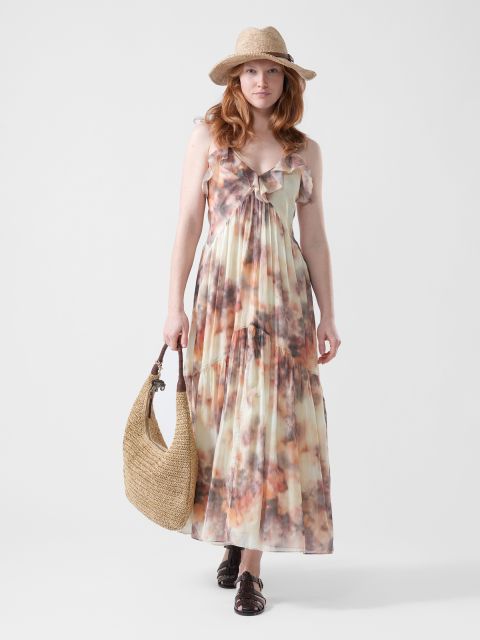 Mujer con un vestido maxi fluido y colorido, sombrero de paja y sandalias, que sostiene un bolso grande tejido en la mano, de pie frente a un fondo blanco liso.