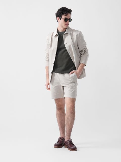 Un hombre con gafas de sol, chaqueta beige, camisa verde oliva, pantalones cortos beige y zapatos marrones se encuentra frente a un fondo blanco liso.