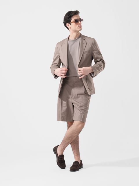Un hombre que lleva gafas de sol, una americana de color topo, pantalones cortos a juego, una camiseta gris y mocasines marrones, posa frente a un fondo blanco liso.