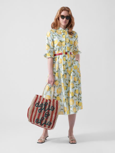 Mujer con un vestido de estampado de limones, gafas de sol y sandalias, sostiene un bolso a rayas rojas y blancas con letras, frente a un fondo blanco liso.