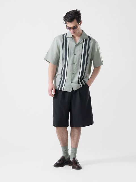 Un hombre que lleva gafas de sol, una camisa de manga corta de color verde claro con rayas verticales negras, pantalones cortos negros, calcetines verdes y zapatos bajos marrones, mira hacia abajo.