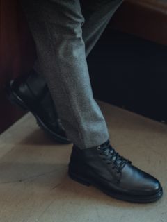 Persona con pantalones grises y botas de cuero negras con cordones, con una pierna cruzada sobre la otra, de pie en un interior sobre un suelo claro.