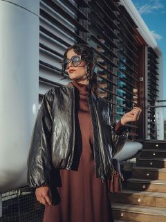 Una mujer con gafas de sol grandes y una chaqueta de cuero oscura se apoya en una pared, con las manos entrelazadas detrás de la cabeza, y posa con confianza a la luz del sol.