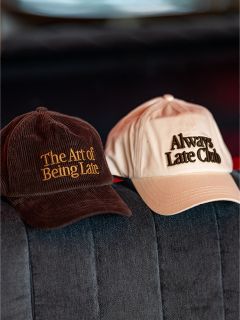 En un sofá gris hay dos gorras de béisbol; en la marrón pone "The Art of Being Late" y en la beige "Always Late Club".