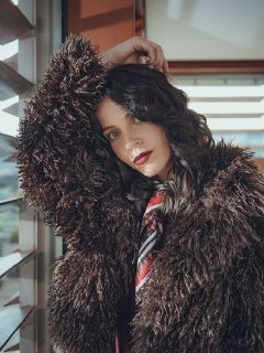 Una mujer con cabello oscuro ondulado lleva un abrigo marrón amplio y esponjoso y una bufanda estampada, y se apoya en una ventana. Mira a la cámara con el brazo levantado mientras la luz del sol entra por las persianas detrás de ella.