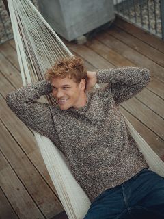 Un joven con cabello rubio rizado, con un suéter marrón y jeans, se relaja en una hamaca, sonriendo y mirando hacia un lado. La hamaca está en una plataforma de madera con una barandilla de metal al fondo.