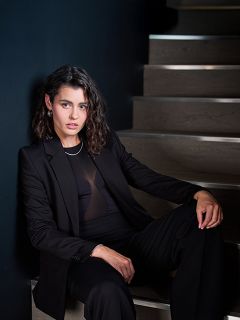 Una mujer con un traje negro está sentada en una escalera de madera frente a una pared oscura y mira con confianza a la cámara, su cabello está peinado en ondas sueltas.