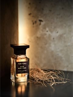 Un frasco del perfume Tom Ford Bois Marocain está sobre una superficie oscura junto a hierba seca, con un fondo cálido y suavemente iluminado.
