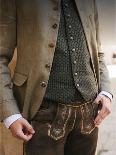 Una persona que viste ropa tradicional bávara, que incluye un chaleco estampado, una chaqueta abotonada y unos pantalones cortos de cuero marrones bordados (Lederhosen).
