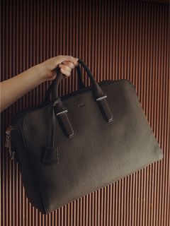 Una mano sostiene un maletín de cuero negro grande con dos asas frente a un fondo marrón acanalado.