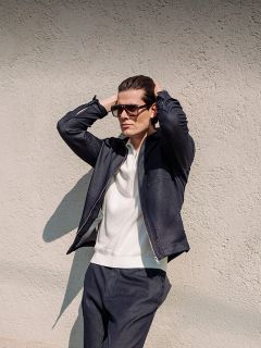 Un hombre con gafas de sol, chaqueta oscura y pantalones oscuros está de pie junto a una pared estructurada, con las manos detrás de la cabeza, mirando a la distancia.