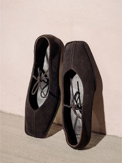 Un par de zapatos de gamuza negros y cuadrados con cordones, de pie frente a una pared y un suelo de color claro.
