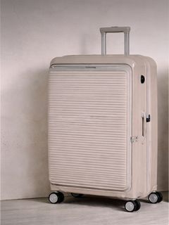Maleta rígida de color beige con ranuras horizontales, de pie sobre cuatro ruedas, con asa telescópica extendida.