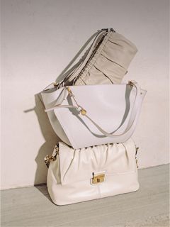 Tres elegantes bolsos de mano color crema están apilados contra una pared clara, mostrando diferentes texturas y detalles de herrajes dorados bajo una suave luz natural.