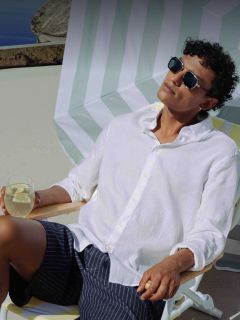 Un hombre con gafas de sol y camisa blanca se relaja en una tumbona, con una bebida con limón en la mano, bajo una sombrilla a rayas en un día soleado.