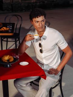 Un hombre con camisa blanca está sentado a una mesa roja, bebiendo un espresso, con un cruasán y un vaso de agua frente a él en un café al aire libre.
