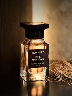 Un frasco de perfume «Bois Marocain» de Tom Ford está sobre una superficie oscura junto a hierba seca, mientras una luz cálida incide sobre él desde atrás.