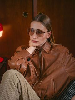 Una mujer con una chaqueta de cuero marrón y gafas de sol está sentada en el interior, con la barbilla apoyada en la mano, junto a una lámpara roja y un armario de madera.