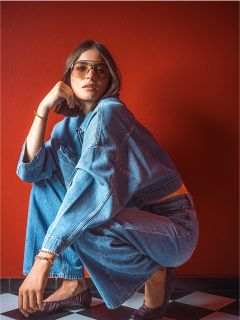 Una mujer con gafas de sol y chaqueta vaquera se agacha sobre un suelo de cuadros blancos y negros delante de una pared roja.
