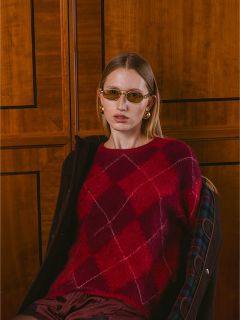 Una mujer con un suéter rojo de argyle, gafas de sol y aretes dorados está sentada frente a una pared con paneles de madera y mira a la cámara.