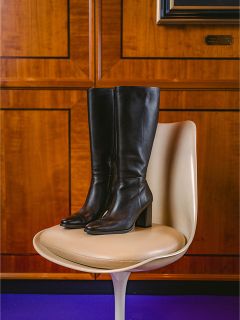 Un par de botas de cuero negras hasta la rodilla con tacón de bloque están sobre una silla moderna de color crema frente a una pared con paneles de madera.