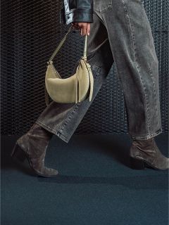 Una persona con vaqueros grises y botas de ante con tacón sostiene un bolso de mano beige y camina frente a un fondo oscuro texturizado.