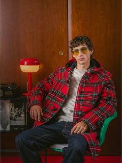 Un hombre con un abrigo de cuadros rojos y gafas de sol amarillas está sentado en una silla verde, con una lámpara roja y una pared de madera al fondo.