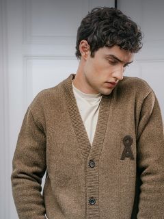 Un hombre lleva una chaqueta de punto marrón con botones y un emblema oscuro bordado en el pecho sobre una camiseta color crema.