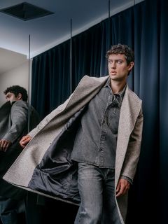 Un hombre con una camisa gris con cuello y jeans se pone un abrigo largo de lana de color neutro.